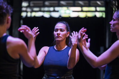 Drie dansers in zwarte kleding, met gefocuste gezichten, voeren een synchroniseerde dansbeweging uit op een donkere achtergrond.