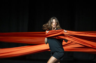 Vrouw in zwarte kleding danst met levendige oranje stoffen in een donkere ruimte.