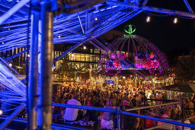 Drukke festivalomgeving met kleurrijke verlichting en een grote versierde tent, omringd door mensen.