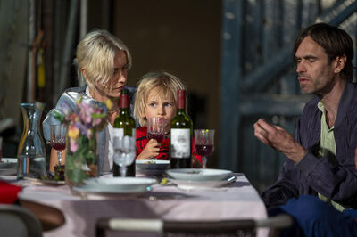 Een man en een vrouw zitten aan tafel met een kind tussen hen, omringd door glazen en flessen wijn.