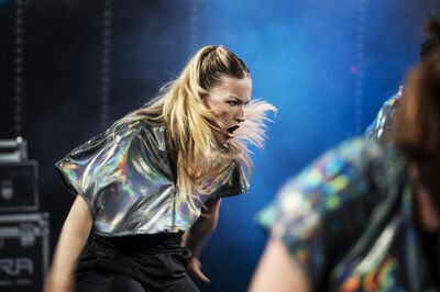 Danseres met lang haar in een glinsterend shirt voert een energieke dans uit op een kleurrijk podium.