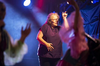 Vrouw danst op een podium met kleurrijke lichten en andere dansers op de achtergrond.