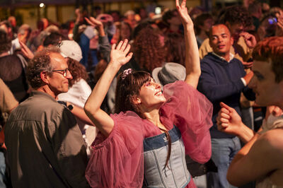 Mensen dansen op een levendig evenement, met één persoon in het middelpunt die vrolijk haar handen in de lucht steekt.