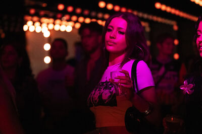 Een vrouw met een drankje staat in een drukke, sfeervolle ruimte met gedimde lichten en aandachtige mensen om haar heen.