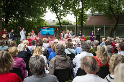 Een menigte zit op stoelen en kijkt naar een optreden in een groene buitenomgeving met acteurs op het podium.
