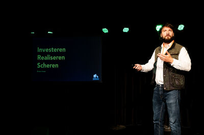 Een man met een witte blouse en groen vest geeft een presentatie.