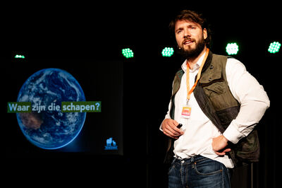 Een man met een witte blouse en groen vest geeft een presentatie.