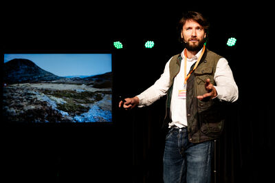 Een man met een witte blouse en groen vest geeft een presentatie.