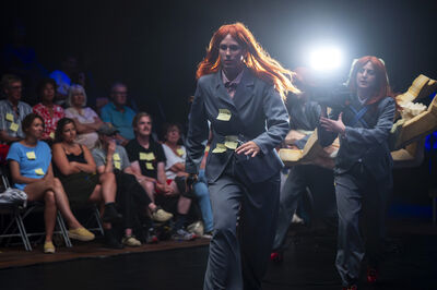 Twee mensen met een rode pruik lopen op een catwalk. Er zit veel publiek naast.
