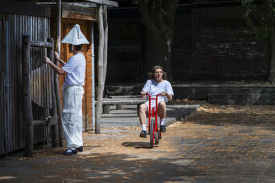Een persoon rijd op een rode kinderfiets en een persoon speelt met drumstokken op houten planken in een speeltuin.