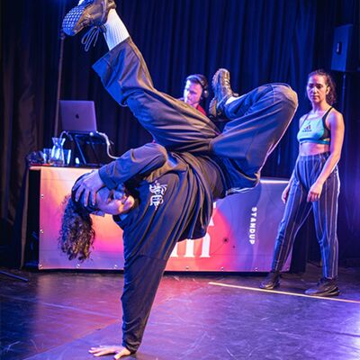 Een vrouw doet een breakdance pose, waarbij ze op een gestrekte arm balanceert en een been buigt. De andere hand bedekt haar gezicht.