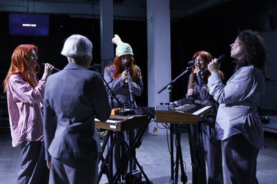 Vijf muzikanten staan in een cirkel rondom keyboards en zingen allemaal in een microfoon.