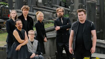 Zeven personen staan en zitten tussen grafstenen op een begraafplaats. Ze dragen voornamelijk zwarte kleding.