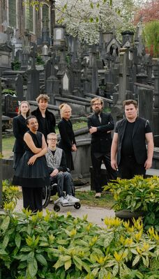 Zeven personen staan en zitten tussen grafstenen op een begraafplaats. Ze dragen voornamelijk zwarte kleding.