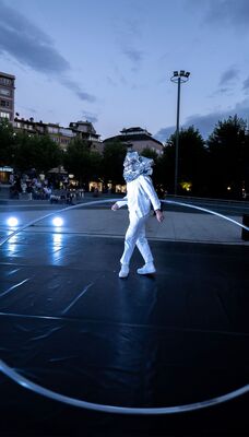 Een circusartiest houdt aan een kant een grote ijzeren hoepel vast. Hun hoofd is volledig bedekt door een zilveren muts.