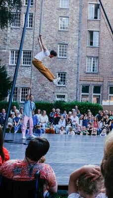 Een acrobaat zweeft hoog in de lucht aan een trapeze tijdens een buitenvoorstelling, terwijl een groot publiek toekijkt.