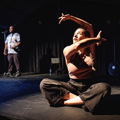 Een danser zit in kleermakerszit met de armen artistiek om het hoofd heen gehouden.
