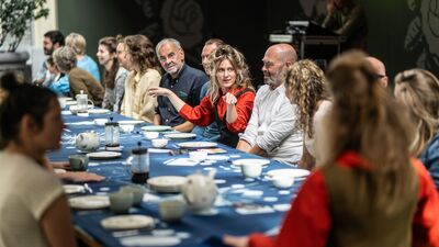 Een grote groep mensen zit aan een lange ontbijttafel. Een vrouw die in het midden zit steekt haar armen uit.