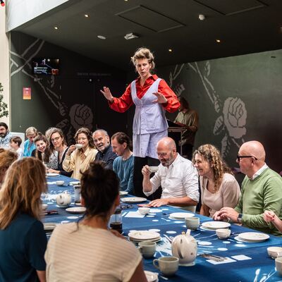 Een grote groep mensen zit aan een lange ontbijttafel. Een vrouw in het midden staat op haar stoel en steekt haar armen uit.