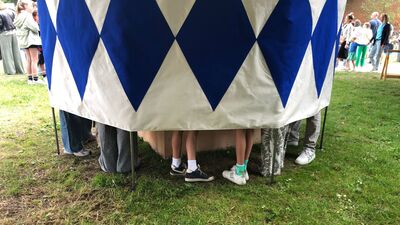 Alleen de benen en voeten van verschillende mensen zijn zichtbaar onder de rand van een rond, wit-blauw geruit zeil van een installatie op een grasveld.