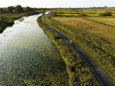 Een luchtfoto van een natuurgebied met een water, fietspad en weilanden.