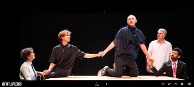 Vijf acteurs staan en zitten rond een tafel in een dramatische scène, waarbij één acteur op de tafel knielt.