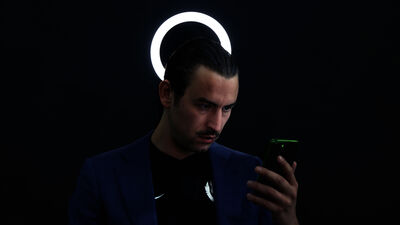 Een persoon staat in een donkere ruimte en kijkt naar een telefoon in zijn hand. Achter zijn hoofd is een ronde witte lamp.