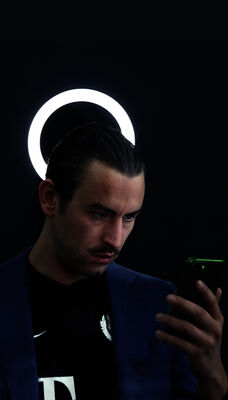 Een persoon staat in een donkere ruimte en kijkt naar een telefoon in zijn hand. Achter zijn hoofd is een ronde witte lamp.