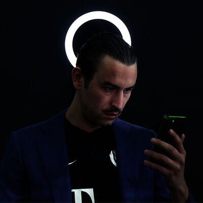 Een persoon staat in een donkere ruimte en kijkt naar een telefoon in zijn hand. Achter zijn hoofd is een ronde witte lamp.