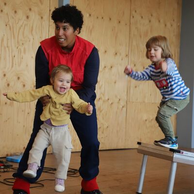 Een foto van een vrouw die een kind optilt met daarnaast een ander kind die op een schuine tafel staat.