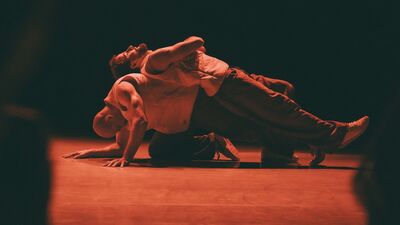 Een danser zit op voorovergebogen op de vloer, terwijl een andere danser rug-aan-rug op de eerste danser ligt.