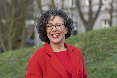 Vrouw met krullend haar in een rode blazer, lachend voor een groene achtergrond met bomen.