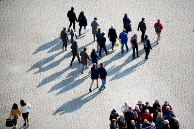 Luchtbeeld van een groep mensen die over een plein lopen, met lange schaduwen op de grond.