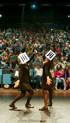 Twee mensen staan tegenover elkaar voor een groot publiek. Hun hoofden zijn bedekt door twee witte blokjes met drie keer de letter "j".