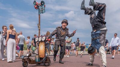 Een circusdirecteur wijst naar een acrobaat die een handstand doet op een zonnige Boulevard. Beiden dragen vintage circuskleding.