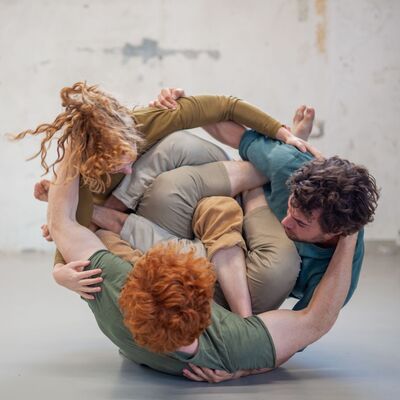 Drie circusartiesten hebben hun benen verworven en houden elkaar vast. In deze houding rollen ze zijwaarts over de vloer.