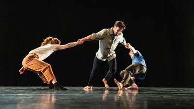 Drie mensen op een donker podium: een centrale danser houdt twee anderen vast terwijl zij naar buiten leunen.