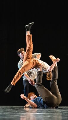 Drie artiesten op een donke podium. Een artiest ligt op de vloer en balanceert een andere artiest met de voet in de rug.