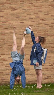 Een persoon doet een handstand en balanceert een kopje op een voet. Iamand anders schenkt thee in dit kopje.