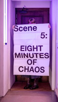 Een vrouw houdt een groot vel papier vast waarop staat: "Scene 5: Eight Minutes of Chaos".