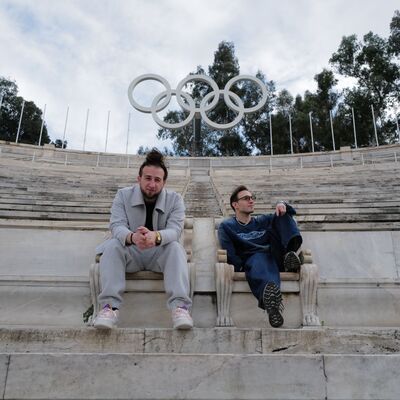 Twee mensen zitten op een verlaten tribune. Het Olympisch logo staat achter hen.