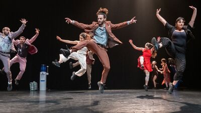 Zeven dansers springen op een donker podium in de lucht.