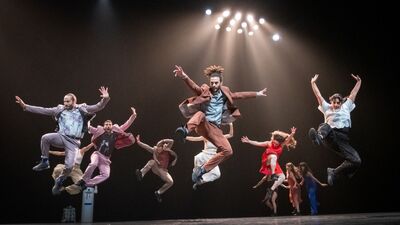 Acht dansers springen op een donker podium in de lucht.