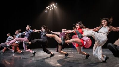 Acht dansers vormen naast elkaar een rij met de armen op elkaars schouders. Ze gooien een been in de lucht.