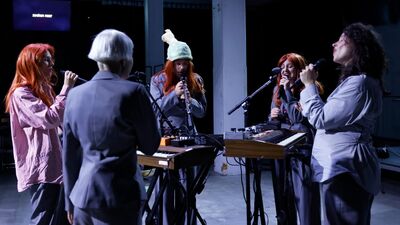 Vijf muzikanten staan in een cirkel rondom keyboards en zingen allemaal in een microfoon.