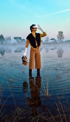 Een persoon met een VR-bril en een hondenmasker in de hand staat in het water van een mistig ven tijdens zonsopgang.