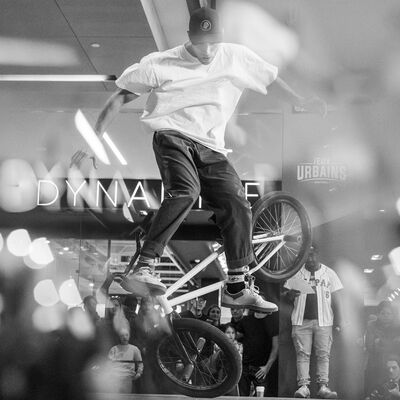 Een persoon met een wit shirt en een pet doet een truc op een bmx fiets.