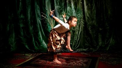 Een jong meisje voert een expressieve dansbeweging uit op een vloerkleed, tegen een achtergrond van een groen gordijn.