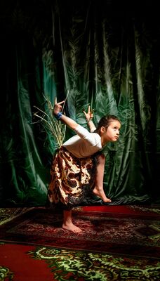 Een jong meisje voert een expressieve dansbeweging uit op een vloerkleed, tegen een achtergrond van een groen gordijn.