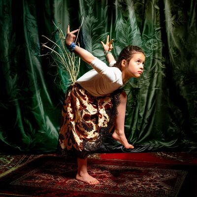 Een jong meisje voert een expressieve dansbeweging uit op een vloerkleed, tegen een achtergrond van een groen gordijn.